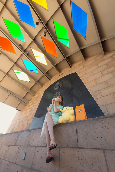 Photo of Lety and the Creature Mastodon stuffed toy sitting on the edge of a stone stage outdoors, directly under a ceiling with colorful glass tiles, during a bright, sunny morning. From top to bottom, Lety is shown wearing a white and mint-green huanengo dress, a white cotton belt, and light tan leather sandals. Her body's facing the camera, her right leg crossed over her left as she uses both hands to hold a white concha to her mouth and looks up at sunlight shining through the diamond-shaped colored glass tiles embedded in the scalloped ceiling.  Sitting to Lety's left is the Creature, looking up at the ceiling, along with an orange takeout bag from La Monarca Bakery, and behind them both is part of “El Niño Perdido” by Alejandro de la Loza.