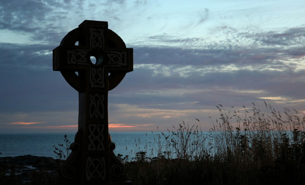 Photograph at daybreak of the silhouette of a Celtic-inspired wooden cross, with a circle around the arms of the cross at the top, situated in tall grass just above a rocky and pebble beach, with the sea behind. The water is a strange shiny blue. The sky is blue but with grey clouds and pink sun rays are getting through at the horizon. 

The cross is just a silhouette in this photo but there is a wooden plaque engraved with the text cited below at the bottom, on the front of its pedestal:
A la mémoire des victimes du naufrage du paquebot Empress of Ireland, 1914
(In memory of the victims of the sinking of the liner Empress of Ireland, 1914)

Of the 1,477 people on board, 1,012 died, making it the worst peacetime maritime disaster in Canadian history. The Celtic cross in Ste-Luce-sur-Mer points towards the wreck, 7 km off the coast.