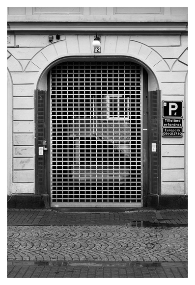 En stående svartvit bild på en port/dörr med gallerdörr neddragen. Huset är av vit sten, under en liten lampa ovanför dörren sitter en liten skylt med husnumret 12. Framför är kullersten och några vattenpölar.