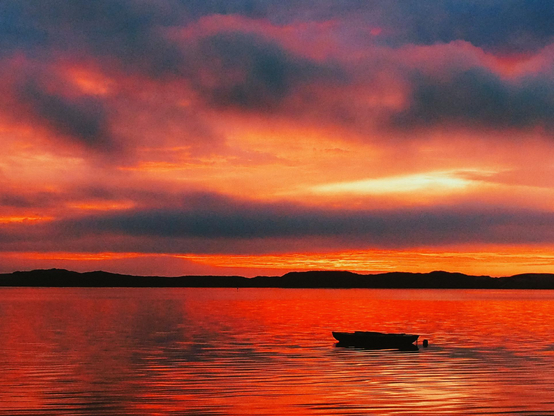 The image shows a landscape with a vibrant sky reflected in calm water. The sky is dominated by shades of orange and red, transitioning to darker hues towards the top of the frame. A single small boat sits on the water's surface, positioned slightly off-center to the right. A dark, silhouetted landmass sits in the background, just above the horizon line. The reflection of the sky is clearly visible in the water, creating a symmetrical and visually striking composition. There is no text visible in the image.