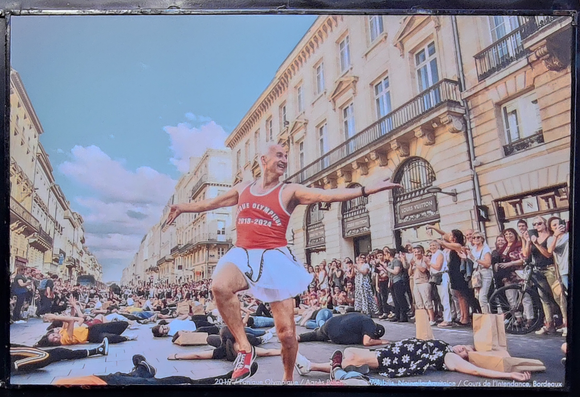 A man in a dance pose on a wide street with crowds on both sides watching, and some people lying on the ground as if injured or worse.