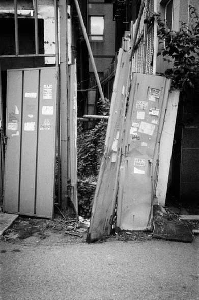 건물을 둘러싼 펜스 중 일부가 무너졌다. 그 안쪽으로 건물 사이로 잡초 따위가 무성하게 자란 것이 보인다.