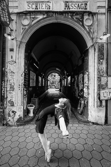 A 12-year-old girl stands on tiptoes in front of an archway labeled 'SCHIERS PASSAGE'. She wears a dark dress over a light long-sleeve shirt. Her body is tilted backward, and her arms hang loosely by her sides. The archway leads to a dark passage with a bright exit at the end. The pavement in front of the arch consists of hexagonal stones. Various posters and graffiti are visible on the sides of the passage.
