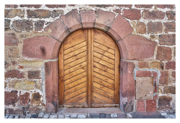 A close-up photograph of an old wooden double door set into a rustic stone wall. The door is arched at the top, framed by a rounded arch made of reddish-pink sandstone blocks. The door panels are constructed from vertical wooden planks arranged in a chevron or herringbone pattern, pointing inward toward the center seam where the two halves meet. The wood appears weathered, with a warm brown tone and visible grain. Surrounding the door, the wall is composed of irregularly shaped stones in shades of red, brown, beige, and gray, mortared together with some white plaster visible in the joints. To the right of the door, there's a small rectangular stone slab or cover embedded in the wall, possibly a access panel or old mailbox slot. The ground in front is paved with uneven cobblestones. The overall scene evokes a historical or medieval European building, with signs of age like dirt, cracks, and slight discoloration on the stones and wood.
