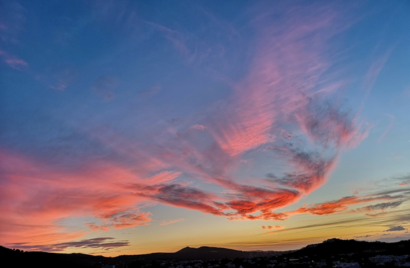 
Das Bild zeigt einen farbenprächtigen Himmel bei Sonnenuntergang. Im oberen Bereich des Bildes ist der Himmel in einem tiefen Blau gehalten. Darunter erstrecken sich breite, langgezogene Wolken, die in leuchtenden Rosa- und Rottönen gefärbt sind. Unterhalb der Wolken geht der Himmel in ein sanftes Gelb und Orange über. Am unteren Bildrand ist eine dunkle Silhouette von Hügeln und Gebäuden zu erkennen.