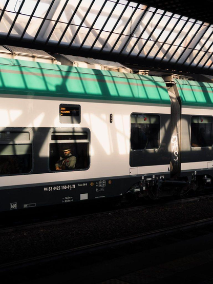 A man with a hat is sitting inside a commuter train at a station. The sun projects light and shadow patterns, and the man is illuminated while the rest of the window is dark.