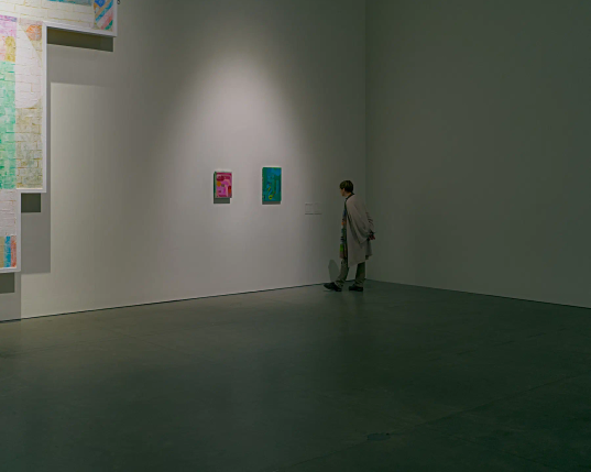 In einem hohen, leeren Raum sind drei Kunstwerke an einer weißen Wand angebracht. 
Links hängt ein beinahe wandhohes, abstraktes Gemälde, das nur zu einem kleinen Teil zu sehen ist. Daneben hängen zwei abstrakte Bilder in Rot und Blau, die kaum größer als ein Blatt Papier sind. Eine Frau steht vor den beiden kleinen Bildern und betrachtet die zugehörige Beschreibung. Die leere rechte Wand und der leere Vordergrund verdeutlichen die Größe des Raumes, in dem sich die kleinen Kunstwerke und ihre Betrachterin beinahe verlieren. 
. 
Three works of art are mounted on a white wall in a high, empty room. 
On the left hangs an abstract painting that is almost as tall as the wall, only a small part of which is visible. Next to it hang two abstract paintings in red and blue, barely larger than a sheet of paper. A woman stands in front of the two small pictures and looks at the associated description. The empty right-hand wall and the empty foreground underline the size of the room, in which the small works of art and their viewer are almost lost. 
. 