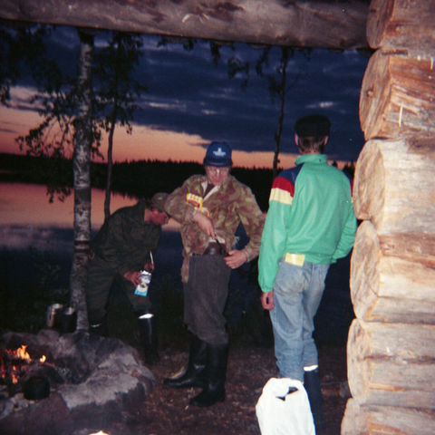 A photo taken from inside a wooden lean-to shelter. Three people in outdoor attire stand near a campfire by a calm lake at dusk.