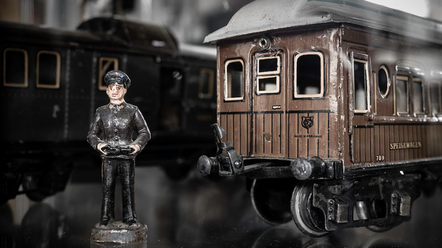 Auf dem Foto rechts sieht man eine Teilansicht von einem alten Peronenwagen der Firma Märklin. Links daneben steht eine kleine Person sie soll wohl einen Schaffner darstellen. Im Hintergrund sieht man noch eine Teilansicht von einem Eisenbahnwagen. Alles steht auf einer Glasplatte.