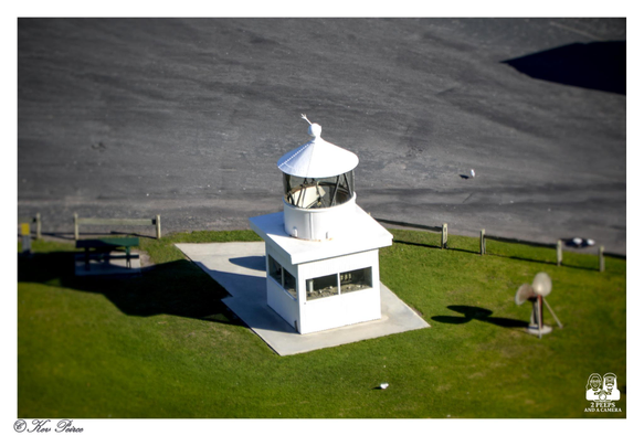 An overhead, slightly tilted photograph of a small, square, white beacon structure with a conical roof and glass lantern room, set on a grey concrete pad.

The beacon is surrounded by bright green grass. In the upper half of the image, there's a dark grey/black paved area. 

A wooden bench and table are visible to the left, and a small, weathered propeller or wind machine sits on the grass to the right.