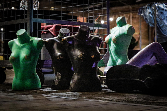 On the photo there are several headless mannequins arranged on the ground in a dimly lit, industrial area. Some are painted green and others black and purple, with chipped and worn surfaces. Most are torso-only figures, positioned upright or lying down near a metal cart and wire fence, under artificial lighting.