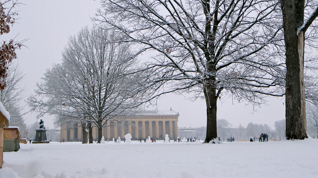 The image shows a snowy landscape featuring a full-scale replica of the Parthenon in the background. The foreground is covered in snow, with bare trees scattered throughout, their branches heavy with snow.
In the distance, there are small groups of people traveling on a walkway to the side of the building, while others build snowmen in an open space in front of it. The sky above is overcast and gray, contributing to the wintry atmosphere of the scene.