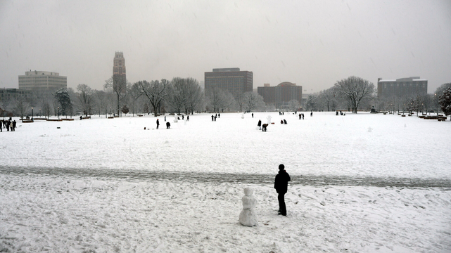 The image depicts an outdoor winter scene covered in snow. In the foreground, there's a small snowman standing on a snowy field. A person is standing near it.
The background features several buildings peeking over the line of leafless trees, including the Nicholas S. Zeppos College tower at Vanderbilt University, as well as campus, retail, and residential buildings.
The sky above is overcast with a grayish hue.