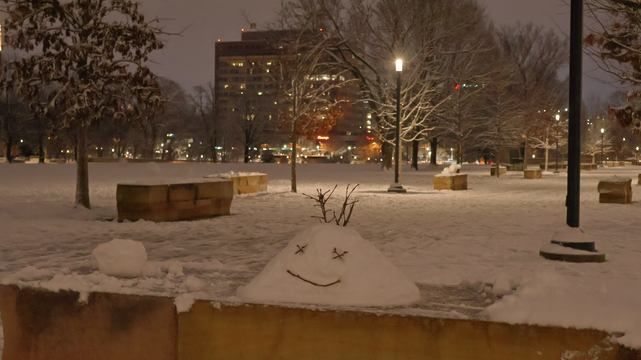 The image depicts an outdoor winter scene in the early morning, as indicated by the dim lighting and the dark sky. The ground is covered in snow, creating a serene white blanket over the landscape.
In the foreground, there are several low stone benches partially buried under snow, with some of their tops visible above the snowline. A small mound of snow sits on top of one bench near the center of the image, and it has been shaped into a simple head sculpture, with facial features defined by small sticks.
The background features leafless trees scattered across the area, their branches bare against the winter sky. In the distance, there is a multi-story building with illuminated windows, suggesting that it might be an office or institutional structure. The overall atmosphere conveys a quiet and chilly urban park setting after a snowfall.