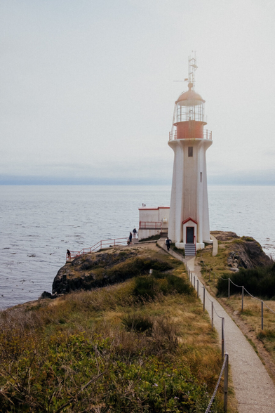 A tall white lighthouse with a red top stands on the edge of a rocky cliff overlooking a calm, grey-blue ocean under a hazy sky. A narrow, winding path bordered by low rope fencing leads from the foreground to the lighthouse entrance. A few people are walking along the path and standing near the lighthouse, admiring the sea view. The surrounding landscape is covered in dry grass, low shrubs, and rugged rocks, adding to the remote, coastal atmosphere. Soft sunlight filters through the mist, giving the scene a serene, slightly nostalgic feeling.