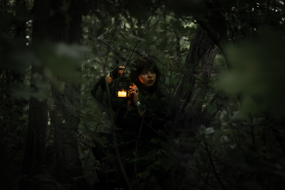 In the photo there's a young woman in a gloomy dark forest. She is holding a lamp with a candle insight. The light is gently elevating the colours as it shifts on her hands and face. She looks focused and slightly mysterious. The numerous shades of green dance around the composition. The vegetation is so thick some leaves obstruct the lens.