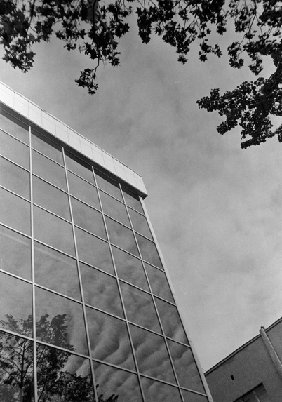 The black and white photo shows a modern building with a wall of many windows. The windows reflect the sky and trees.