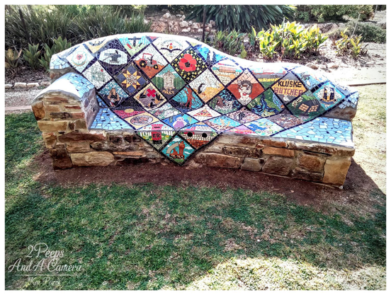 A unique outdoor stone bench, with the seat and backrest completely covered in a colorful mosaic.

The mosaic is arranged in a diamond or quilt pattern, with each tile depicting a different image, seemingly related to local history, trades, or farming, including a tractor, a poppy, an anvil, a pair of boots, and one section with the words "KLUSKE BUTCHER."

The bench sits on a patch of grass in front of a garden bed with lush green and brown foliage.