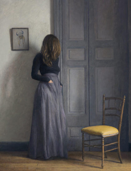 A solitary woman stands near a door, wearing a black top and a long gray skirt. She has her back turned, with her hands in her pockets. Beside her is a wooden chair with a yellow cushion, and a framed artwork is hanging on
