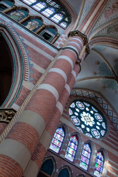 A photograph of the interior of a church, taken diagonally from below. From the left corner to the right is a large pillar, painted with small, brown-red bricks alternating with beige, stone-colored bands. At the top, the pillar flares out, with painted borders that extend into the ceilings to the left and right. To the left of the pillar, the wall above a large arch is also painted with stones. Within it are four closed windows, the middle two of which are painted with saints. Above that, a multicolored stained-glass window. At the top right, first, the ceiling is painted with subdued green motifs, and below that, in the wall, a large, round, multicolored stained-glass window (with floral motifs), and below that, four arched stained-glass windows depicting saints.