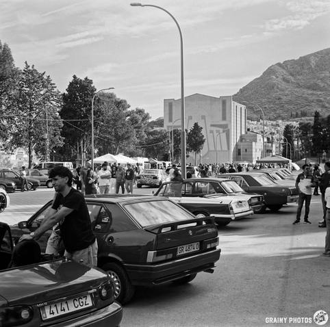 A bustling outdoor car meet featuring rows of vintage cars under a partly cloudy sky. People are interacting and enjoying the event, with mountains in the background and a modern building nearby.