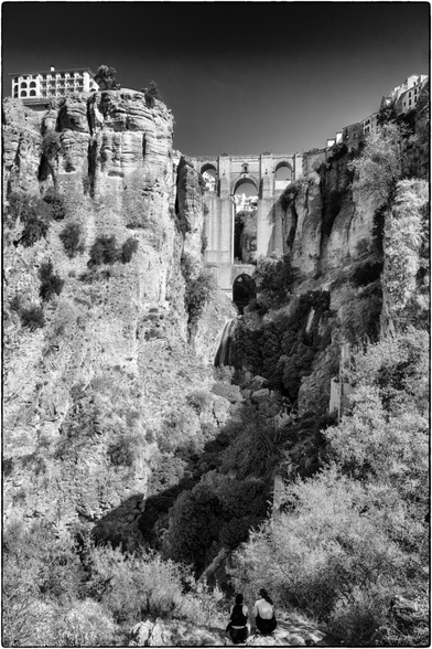 Schwarz-Weiß-Aufnahme einer tiefen, zerklüfteten Schlucht. Hohe, steile Felswände dominieren die Szene. Am oberen Rand überspannt eine imposante, hohe Steinbrücke mit mehreren Bögen die Schlucht, und auf den Klippen dahinter sind Gebäude zu erkennen. Im unteren Bereich der Schlucht, in der Mitte, fällt ein Wasserfall herab. Im Vordergrund, unten am Rand, sitzen zwei Personen mit dem Rücken zum Betrachter auf einem Felsvorsprung und blicken in die Tiefe der Schlucht hinauf zur Brücke.