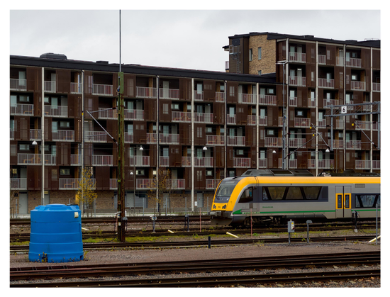 En liggande bild på ett stort flerfamiljshus i brunt med regelbundna lägenheter med växlande bruna och vita staket. I förgrunden järnväg med ett gult och grått tåg. En blå cylindrisk låda till vänster vid järnvägen.