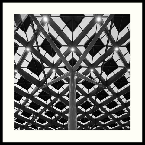 b&w - Square format image that shows a structural pillar and roof of Central Station in The Hague. The shot is taken from an angle and point of view that creates a vertical symmetry. The roof itself is also symmetrical and made of diamond-shaped glass windows placed in a framework of stainless steel.
