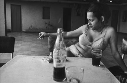 Mujer sentada en una habitación humilde, con una botella de Coca-Cola y un vaso sobre la mesa, fotografía en blanco y negro 