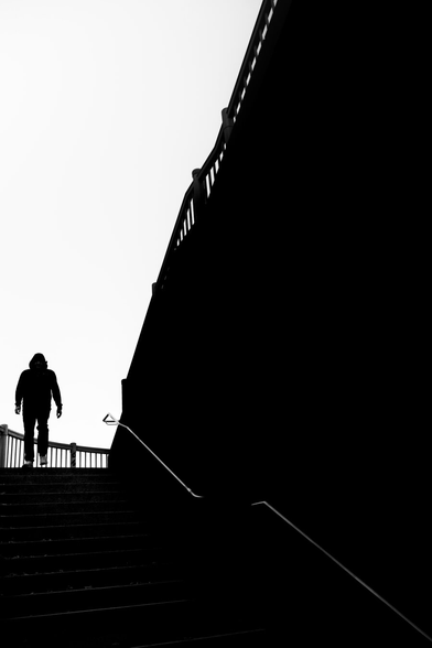 Black and white backlit portrait shot. Urban scene. The silhouette of a man walking down a staircase stands out clearly against the cloudless sky.