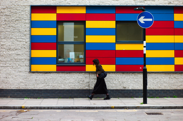 The image showcases a vibrant street scene. The primary focus is a woman walking in profile across a sidewalk. She is wearing a dark, long dress, a dark jacket, and boots, with a black bag slung over her shoulder.

The background is dominated by a building facade. A significant portion of the wall is constructed from light-colored, textured bricks. Above the bricks, there's a section with a highly colorful design composed of rectangular panels. These panels are arranged in a grid pattern and feature alternating colors: red, yellow, and blue. Two windows are visible within this colorful section, their dark frames contrasting with the bright hues.