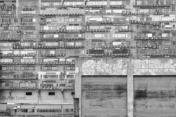 This is a black and white architectural photo of the rear of a mixed use building. Hong Kong (2016).
The entire image is filled with the rear of somewhat ugly building that resembles an old printed circuit board. The lower right corner is filled with two large, perhaps ten metres square, roller shutters. The remainder of the building is made up from thirteen (maybe not considered an unlucky number in Hong Kong?) floors. Each floor is highly random with different style windows, multiple, random spaced external air conditioning units, the only thing missing is drying laundry. 
The location: On the Hung Hom waterfront, Kowloon, Hong Kong.