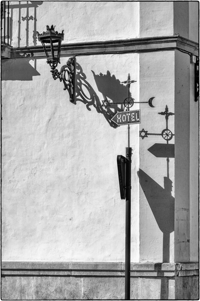 Schwarz-Weiss-Detailaufnahme einer hellen, texturierten Wand eines Gebäudes, vermutlich bei starkem Sonnenschein. Links oben hängt eine reich verzierte, schmiedeeiserne Wandlaterne, deren komplexer Schatten auf die Wand wirft. Rechts davon befindet sich ein Pfosten mit mehreren Schildern, darunter ein rechteckiges Schild mit einem Pfeil und dem Wort "HOTEL". Rechts vom Pfosten sind mehrere kreuzförmige Dekorationen und Symbole (Stern und Mondsichel) befestigt, die ebenfalls lange, scharfe Schatten auf die Wand werfen.