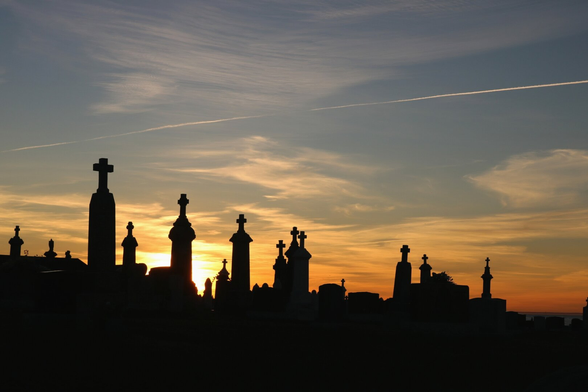 Photograph of the black silhouettes of old tombstones in a cemetery, with the setting sun behind them. against an orange sky on the horizon and blue above, with a few white clouds and the contrail of an airplane. The stones are tall, tower-like, each with a cross on top.

Photographie de la silhouette noire de pierres tombales anciennes dans un cimetière, avec derrière le soleil qui se couche, dans un ciel orange à l'horizon et bleu plus haut, avec quelques nuages blancs et la trace d'un avion. Les pierres sont hautes, en forme de tour, avec chacune une croix sur le dessus.