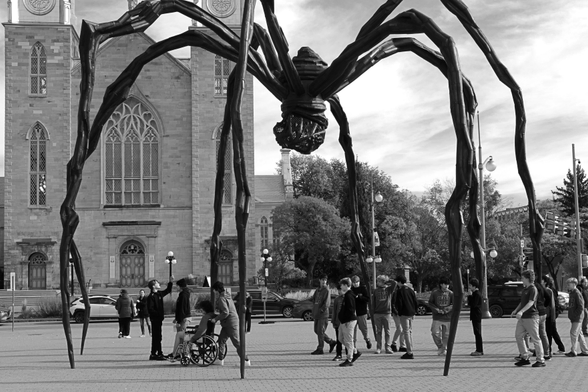 Outside the Gallery, Maman, the large bronze spider sculpture stands. Walking underneath it and between the eight legs is a group of young people including a wheelchair user, smiling and taking pictures.