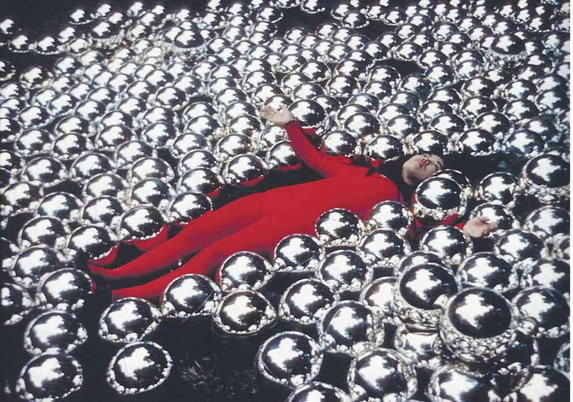 Person in a red jumpsuit lying submerged among numerous reflective silver spheres.
