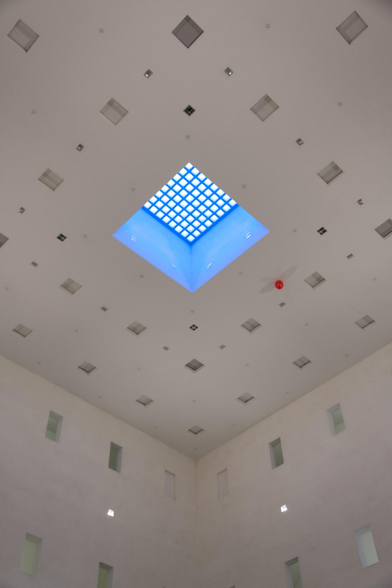 we're looking diagonally towards the ceiling of a large white room. in the center, there is a blue opening with a grid. a lost red balloon hangs under the ceiling. there are many openings and small windows in walls and ceiling. daylight. 