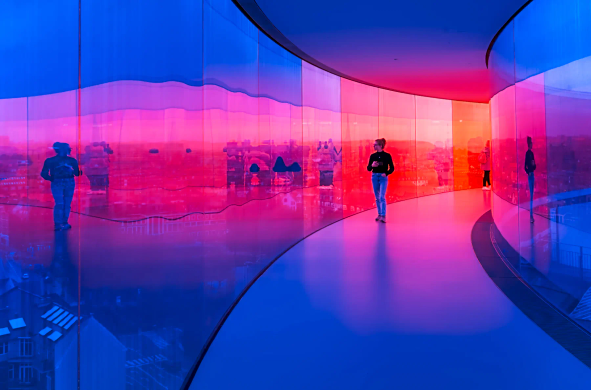 Ein langer runder Gang ist zu beiden Seiten in Glas eingefasst. Im Vordergrund sind die Scheiben noch blau, Richtung Hintergrund wechselt die Farbe langsam zu rot. Im hinteren Drittel des Ganges wandert eine junge Frau mit einer Kaffeetasse in der Hand an den Glasscheiben entlang und betrachtet die Stadt. Vielfältige Spiegelungen verzerren den Blick nach draußen. 
.
A long, circular corridor is enclosed by glass on both sides. In the foreground, the panes are still blue, but towards the background, the colour slowly changes to red. In the rear third of the corridor, a young woman walks along the glass panes with a coffee cup in her hand, looking out at the city. Diverse reflections distort the view outside. 
.
