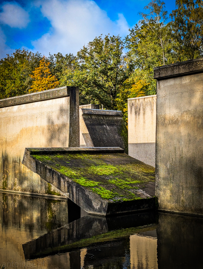 An architectural structure partially submerged in dark water, set against a backdrop of vibrant autumn foliage and a blue, cloudy sky.

A large, flat concrete slab is the central focus, angled dramatically, with its lower edge submerged in the water. The slab's surface is covered in patches of bright green moss, providing a vivid contrast to the muted, earthy tones of the concrete. 

Above the concrete walls, a dense cluster of trees forms the background. The leaves show autumn colors, with deep greens transitioning into bright yellows and oranges, especially on the left.