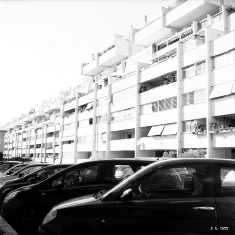 A black and white photograph of a street in a residential area with a row of cars parked in the foreground and a long apartment building in the center. White sky above.