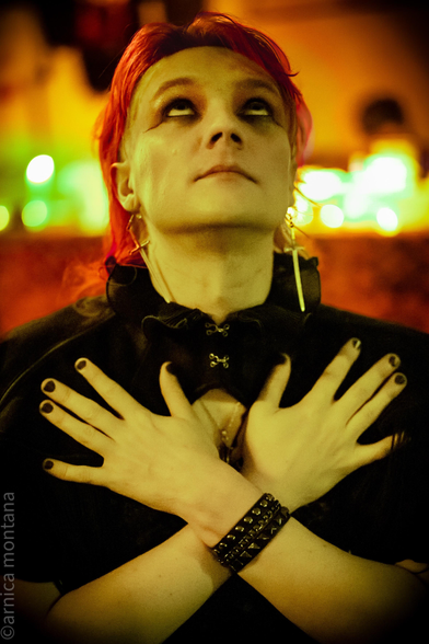 A person with bright red hair, wearing dark makeup and gothic attire, poses with arms crossed against their chest. The background features warm, colorful lights, creating an atmospheric effect.