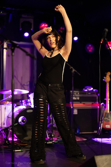 A performer dancing on stage, wearing a black outfit with fishnet sleeves and ripped pants. The background features musical instruments and colorful stage lighting.