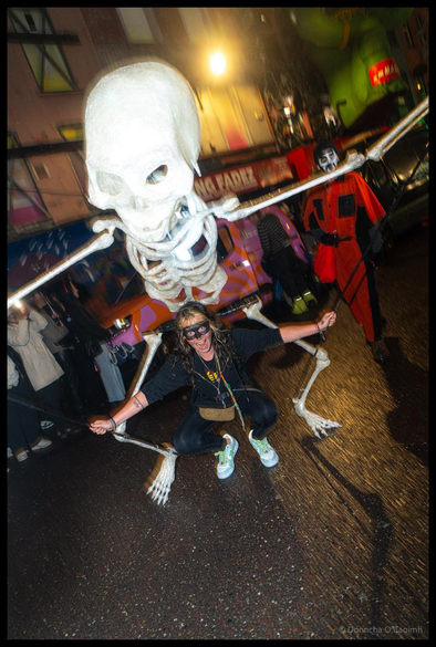 A woman in black clothing and face mask crouches on North Main Street in Cork whilst manipulating a massive white skeleton puppet overhead during the Dragon of Shandon parade at night, with illuminated shop fronts and other costumed participants visible in the background.