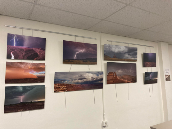 A plain off-white interior wall. Nine medium sized brightly colored, frameless landscape prints are hanging on it. All of the images are storms and lightning bolts over desert landscapes.