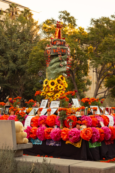 Photo of the Creature Mastodon stuffed toy sitting on a concrete bench in front of a large community ofrenda at a park during sunset. Towards image bottom-left is the Creature, sitting on a bench built into the concrete edging of a garden bed and looking toward the community ofrenda “Let Freedom Ring: Honoring Our Immigrant Ancestors” by Consuelo G. Flores. The multi-level ofrenda is themed after the raised arm and torch of the Statue of Liberty, and decorated with Día de Muertos motifs such as monarch butterflies, cempasúchil flowers, and papel picado. All along the ofrenda are portraits of departed loved ones, placed by residents of the community.