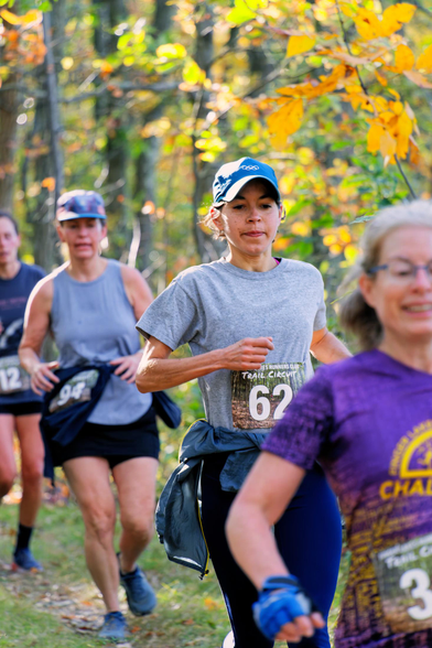 Four women are running across a vertical frame with orange and green leaves and tree trunks in the background
