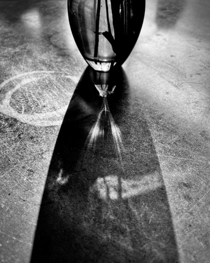 A glass vase casts a long shadow on a textured surface, revealing intricate patterns in black and white light.
