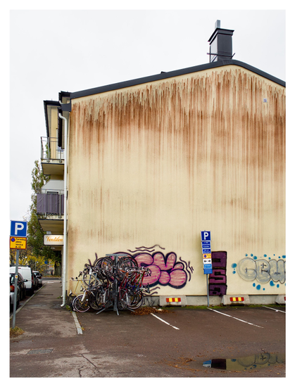 En stående bild på ett tiotal cyklar som hänger runt en stolpe framför en fönsterlös, gul husvägg med röda ränder och lite graffiti. Framför hussidan är en tom bilparkering. Till vänster är bilar parkerade på gatan. På huset vänstersida hänger belyst skylt med texten ”Ventilen cykelverkstad”.