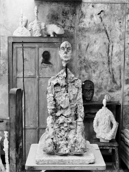 Sculpture d’une figure masculine assise, par Giacometti, dans son atelier, avec d’autres sculpture : un plus petit buste blanc sur la droite est posé sur un tabouret, d’autres bustes sont posés sur une grande armoire au fond à gauche; les murs sont crayonnés.