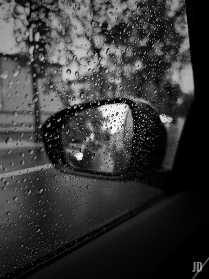 Fotografía en blanco y negro tomada desde el interior de un vehículo durante la lluvia. La ventanilla lateral y el espejo retrovisor exterior están cubiertos por innumerables gotas de lluvia de diversos tamaños, algunas formando pequeños charcos. El espejo, oscuro y central, refleja una escena borrosa con luces difusas. Al fondo, a través de la ventanilla, se distinguen contornos difusos de árboles o vegetación y una calle, todo muy desenfocado. Una porción oscura del interior del coche se ve en la esquina inferior derecha.